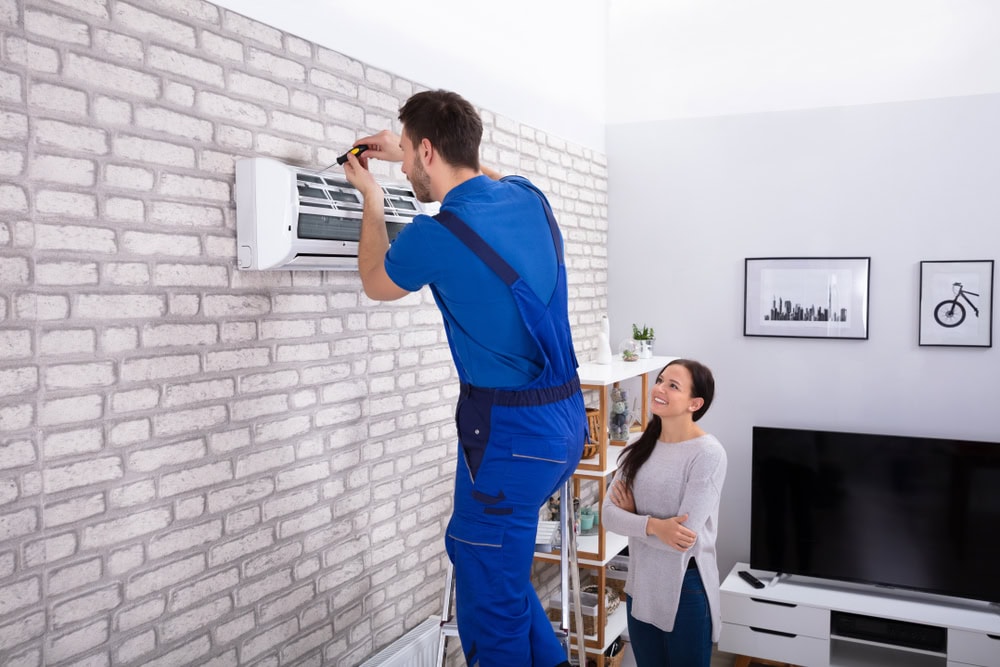 A technician in a blue uniform stands on a ladder, repairing an air conditioner mounted on a brick wall. A woman stands nearby, watching him.