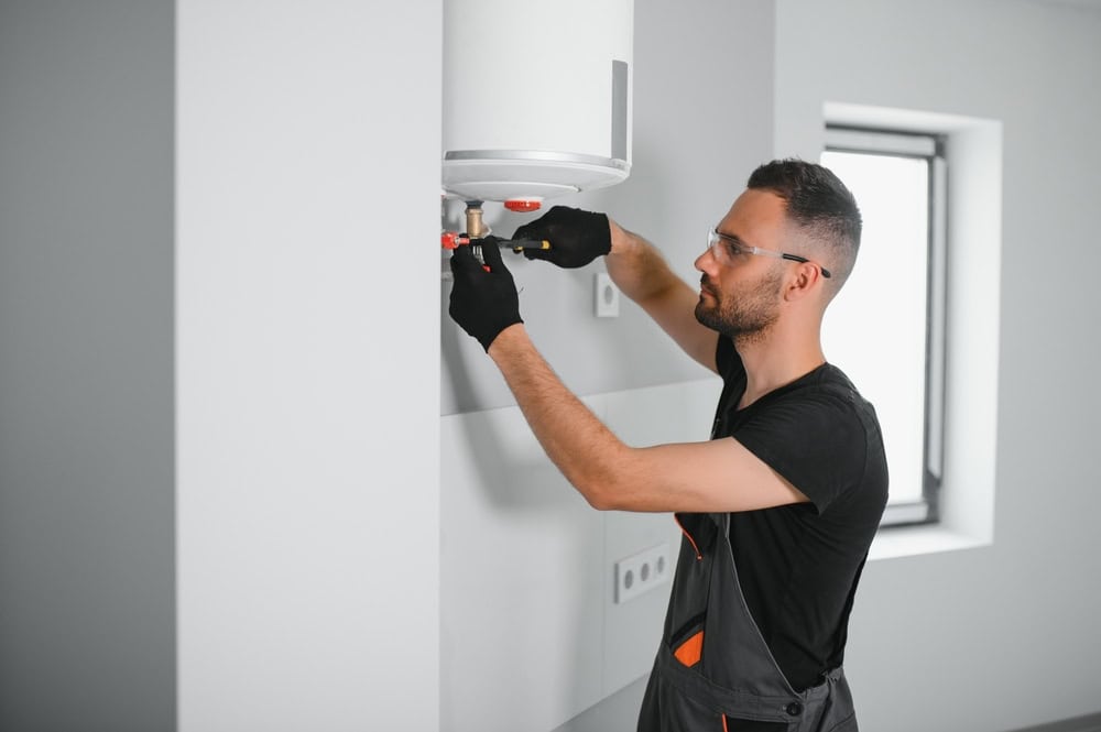 A person wearing protective goggles and gloves is using a tool to adjust a wall-mounted water heater in a bright room.