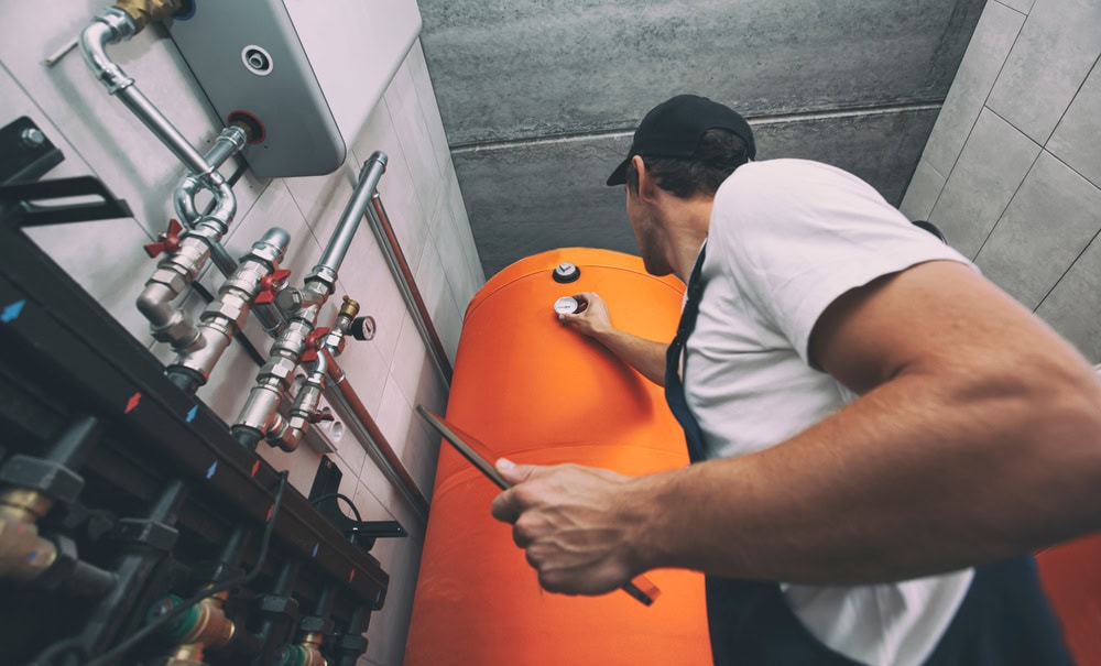 A person wearing a cap and work overalls adjusts a gauge on a large orange industrial tank—showcasing the expertise of an HVAC Contractor North Attleboro—in a utility room filled with pipes and valves.