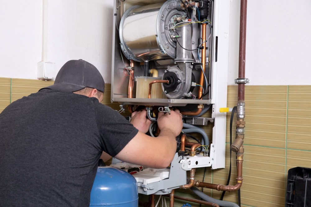 A person wearing a gray cap and t-shirt is repairing or inspecting the inside of a wall-mounted boiler or water heater, surrounded by pipes and wiring in a tiled utility room—expert service like AC Installation North Attleboro, MA.