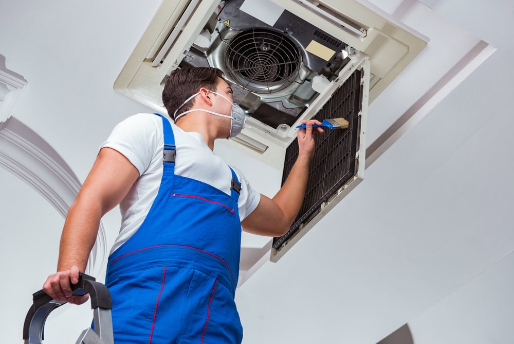 A maintenance worker in a blue uniform is standing on a step ladder and cleaning a ceiling air conditioning unit.
