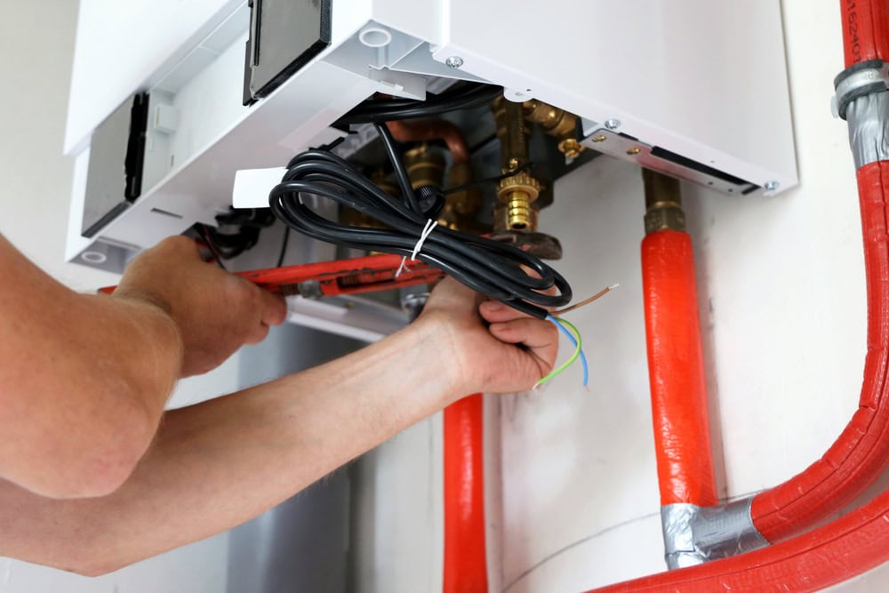A person is using tools to install or repair a wall-mounted boiler. Red pipes are connected to the boiler, and electrical wires are visible.