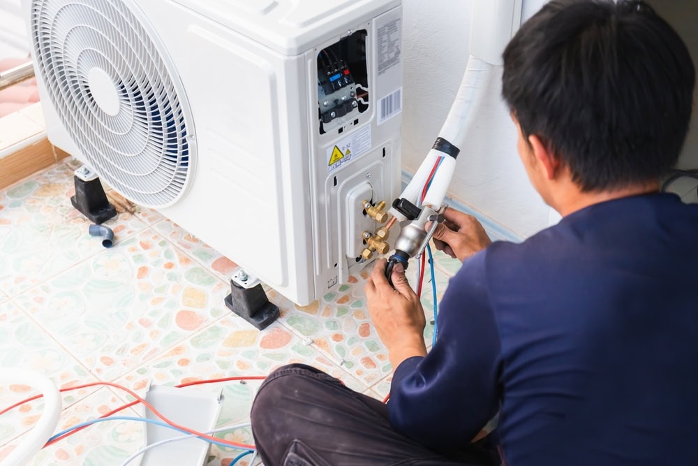 A technician in a blue shirt installs or repairs an air conditioning unit, using tools on the pipes and connections. As part of AC Installation North Attleboro, the unit is mounted on the floor next to a wall inside a room.