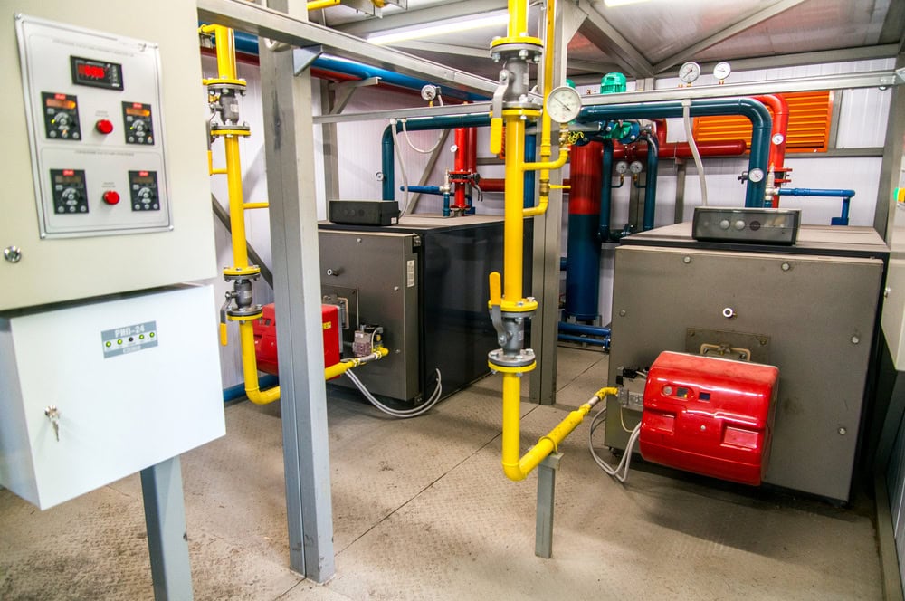 A room containing industrial equipment with two large boilers, colorful pipes, and control panels.