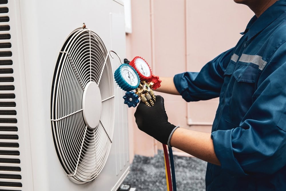 A technician in a blue uniform uses a manifold gauge to check or repair an outdoor air conditioning unit. An HVAC Contractor North Attleboro expert, he adjusts the equipment with one hand while holding the gauge in the other.