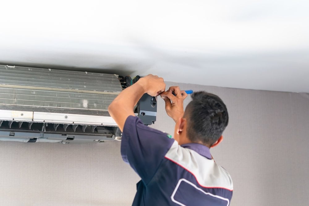 A technician in uniform repairs or installs an air conditioning unit mounted near the ceiling, using a screwdriver and other tools—expert AC Installation North Attleboro residents can rely on.