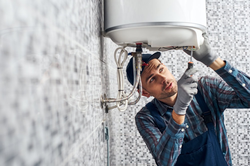 A plumber in gloves and overalls uses a screwdriver to repair a wall-mounted water heater in a tiled bathroom, ensuring quality service you’d expect from an HVAC Contractor North Attleboro, MA residents trust.