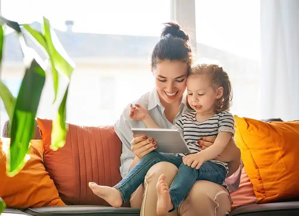 A smiling woman and a young child sit on a sofa, looking at a tablet together. The child sits on the woman's lap. The background includes a window and green plant, with orange and yellow cushions around them.