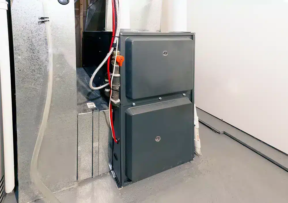 A gray furnace is installed in a basement with two visible access panels. Red and white wires extend from the top, and a clear plastic tube runs along the side. The surrounding area has a concrete floor and plain walls.