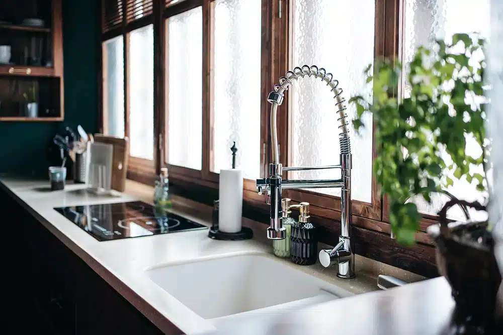 A modern kitchen with a white countertop featuring a deep sink and a tall, flexible faucet. Wooden windows allow natural light in, and a potted plant adds greenery. A stovetop and kitchen utensils are seen in the background.