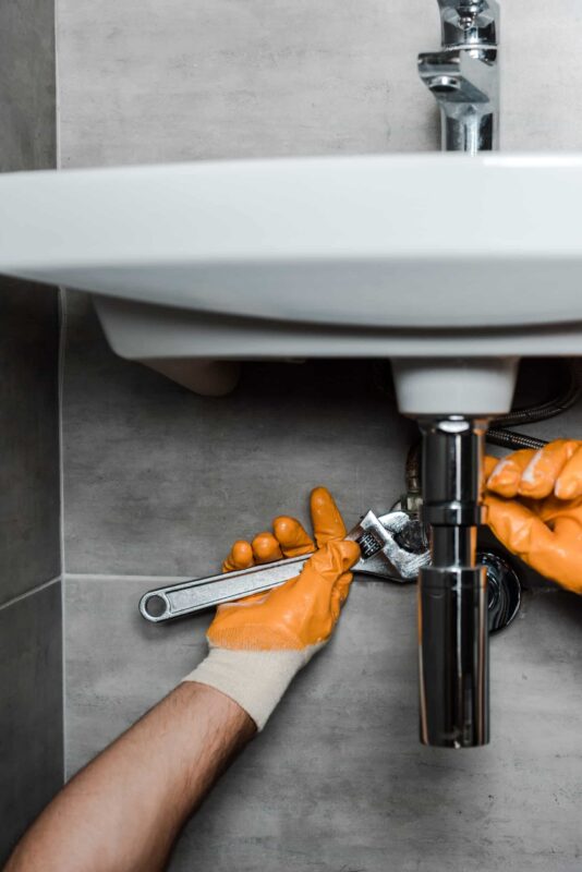 A person wearing orange gloves is using a wrench to fix plumbing under a white bathroom sink. The faucet and part of the tiled wall are visible.