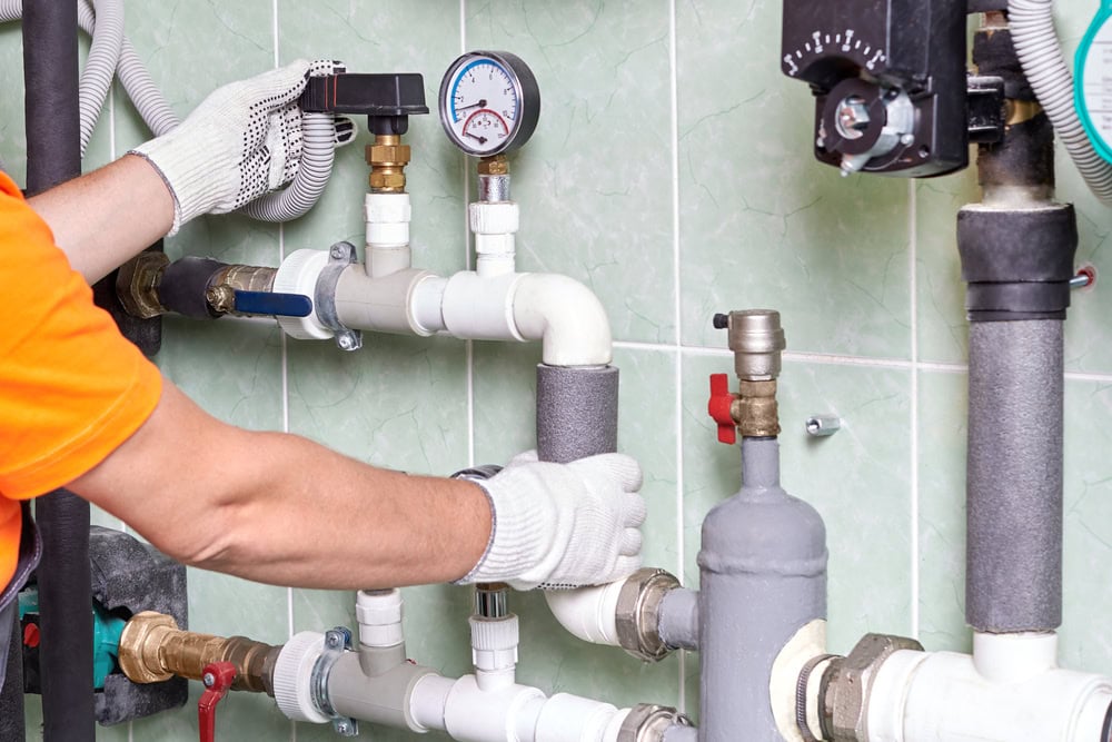 A person wearing gloves and an orange shirt is adjusting gauges and valves on a network of white PVC pipes. The setup is mounted on a green-tiled wall, featuring pressure meters and various fittings.