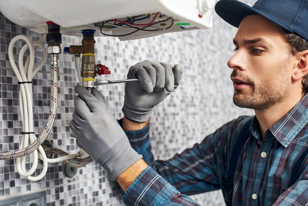 A plumber in a blue cap and plaid shirt uses a wrench to fix pipes under a sink. He is focused on his task, with gloves on for protection. The wall features a gray tile design.