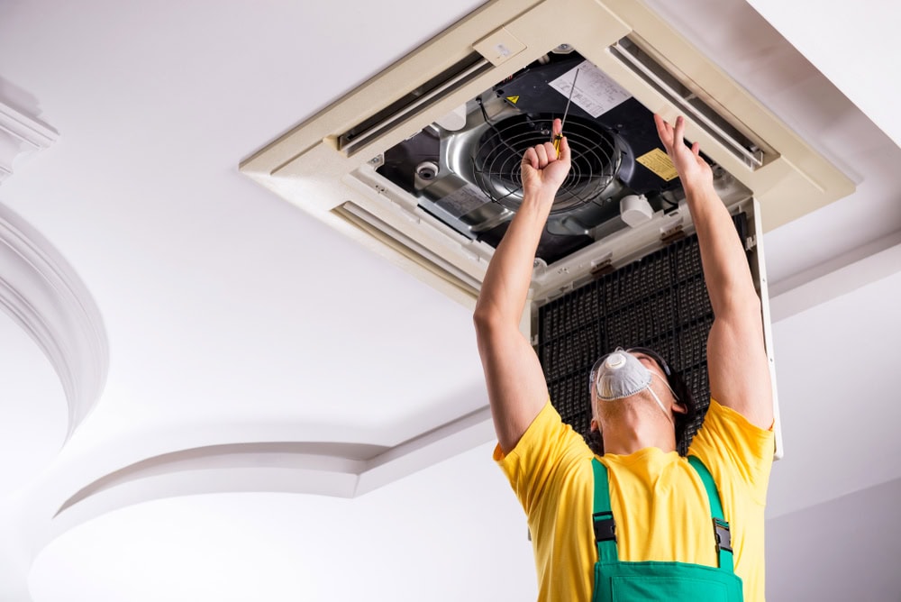 A person wearing a yellow shirt, green overalls, and a protective mask is standing on a ladder, reaching up and repairing a ceiling-mounted air conditioning unit.