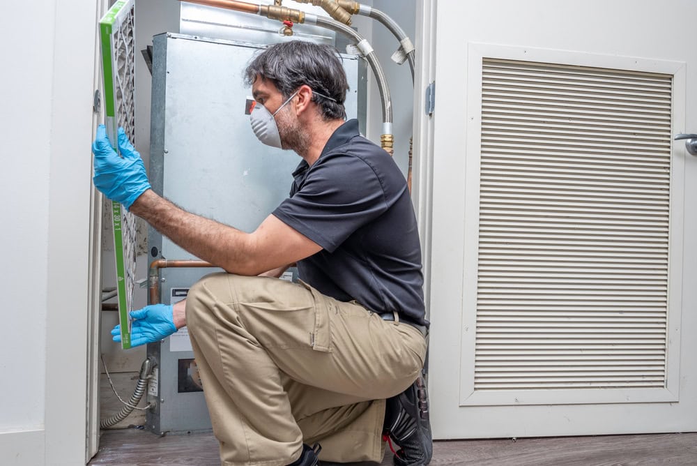 A man wearing a protective mask and gloves kneels while inspecting an HVAC system. He holds a filter in one hand, examining the unit closely. The setting appears to be indoors near a utility closet.