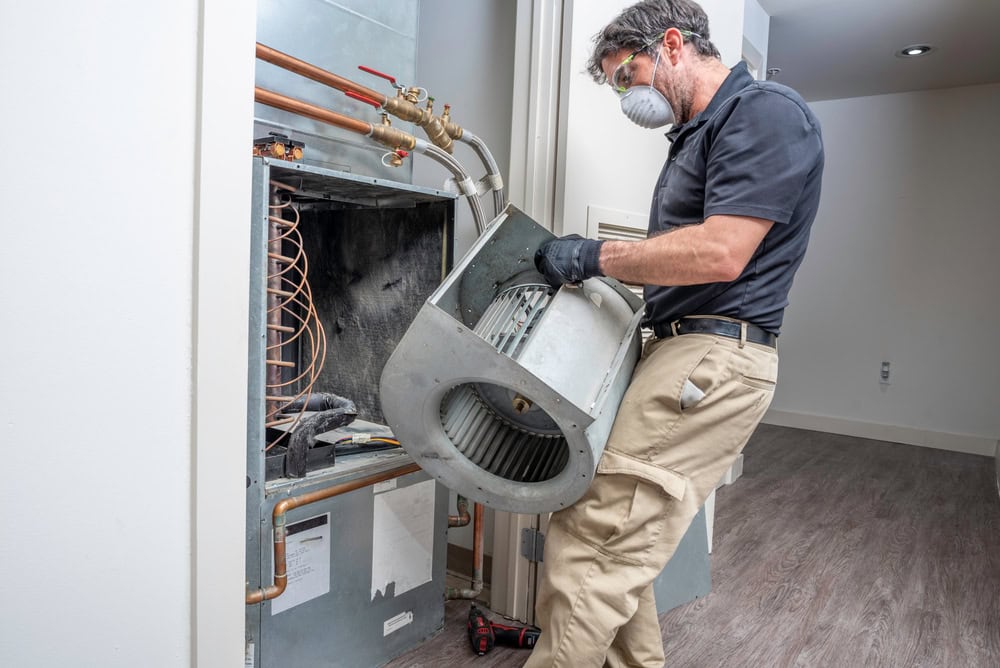 A technician wearing a mask and gloves is handling a large component from an HVAC unit inside a modern room with wooden flooring. Various tools and copper pipes are visible around the open unit.