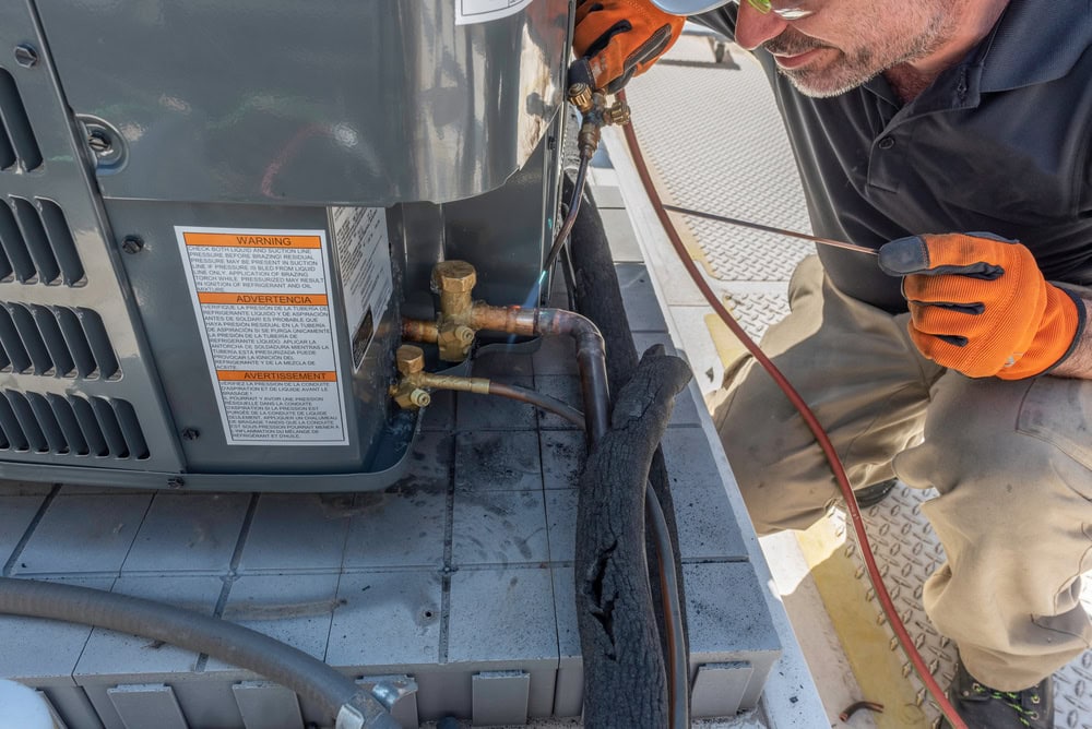 A technician wearing gloves and sunglasses is working on an air conditioning unit. He is adjusting wires and valves with a tool in a mechanical setting. The unit has visible labels and multiple pipes.