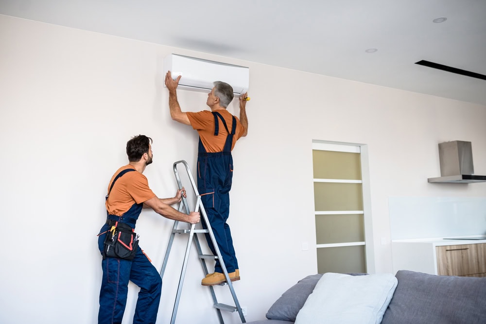 Two technicians in blue overalls and orange shirts install an air conditioning unit on a white wall. One stands on a ladder while the other holds the unit. The room has modern decor with a sofa and kitchen in the background.