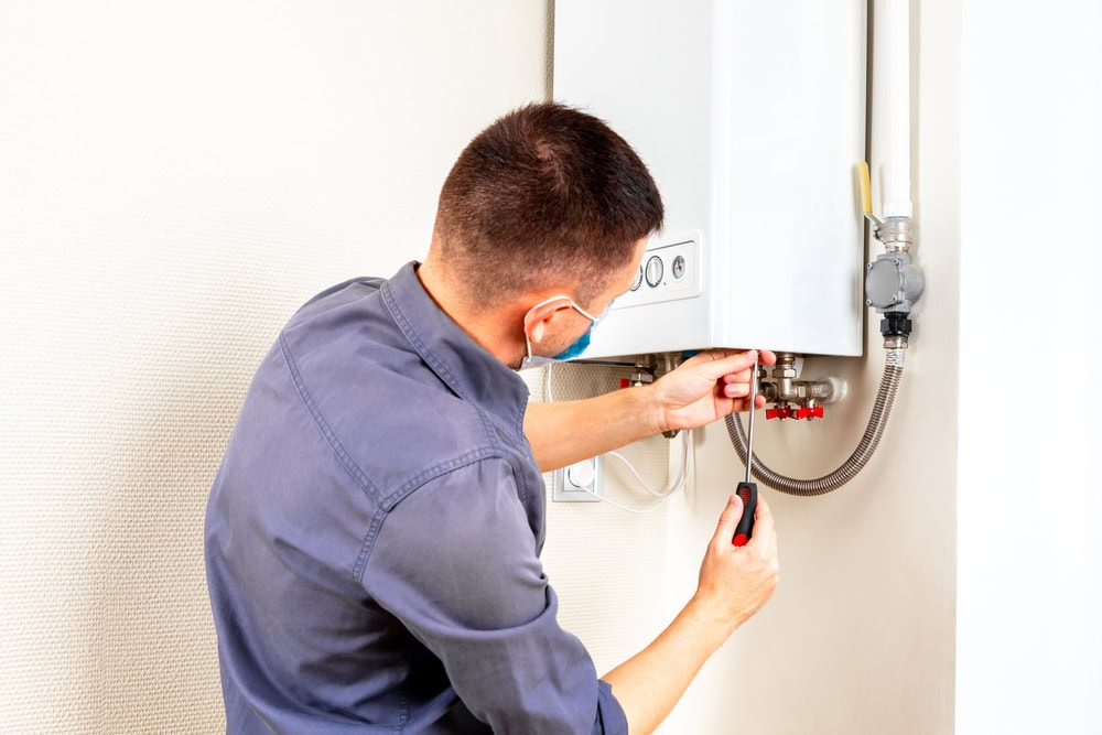 A person wearing a mask and a blue shirt uses a screwdriver to adjust a white gas boiler mounted on a wall. The boiler has knobs and pipes, and they are working carefully with focused attention.
