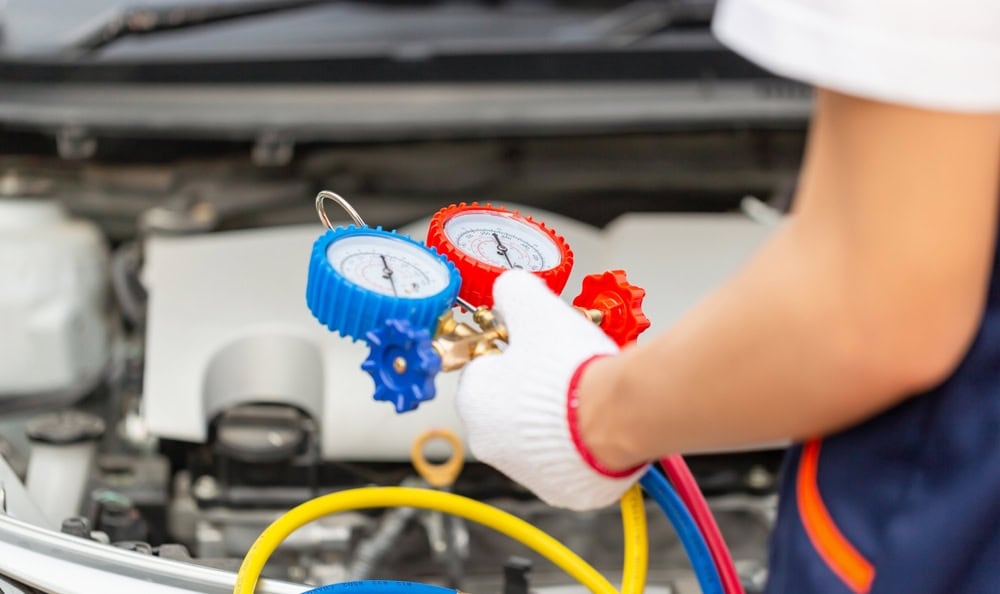 A person wearing gloves uses a dual gauge tool to check a car's air conditioning system under the hood. The tool has a blue and a red dial connected to hoses, indicating pressure readings.