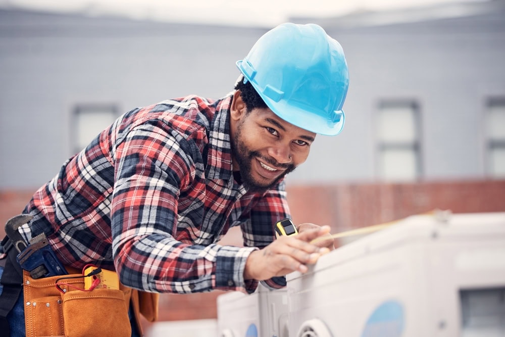 A construction worker wearing a blue hard hat and plaid shirt smiles while using a tape measure to assess building materials. A tool belt is around the worker's waist. The background is a blurred building facade.