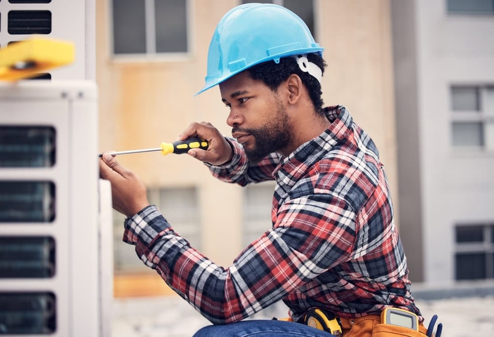 A construction worker in a blue hard hat and plaid shirt uses a screwdriver to fix an outdoor unit, possibly an air conditioner, against a building backdrop. He has a tool belt around his waist.