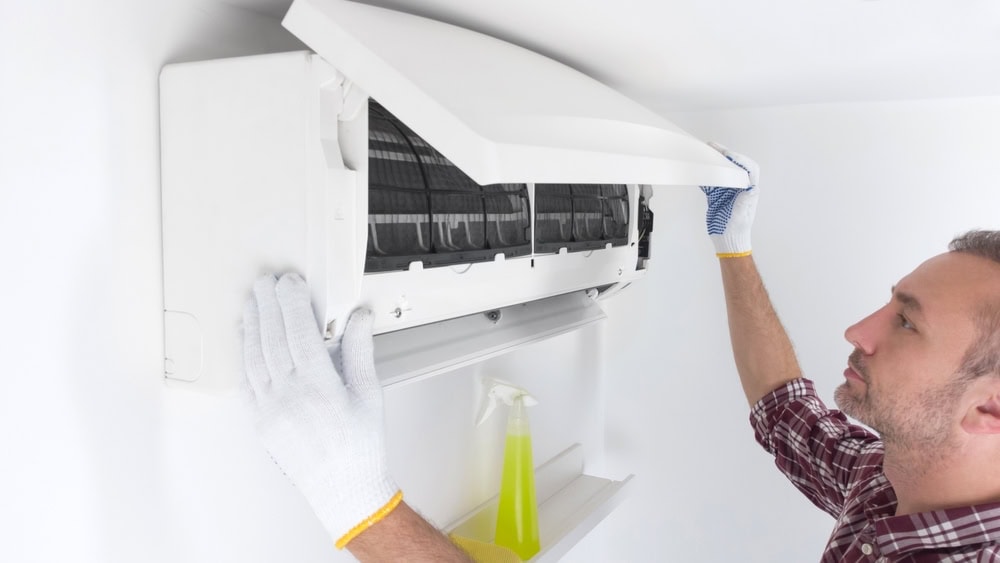 A man wearing a checked shirt and gloves cleans an air conditioner mounted on a wall. He lifts the front cover, revealing the filters inside. A spray bottle and cloth are on a nearby shelf.