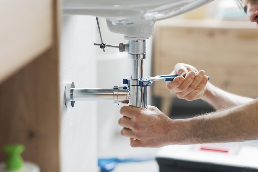 A person uses a wrench to tighten a pipe under a sink in a bathroom. The metal pipe connections are shiny, and the scene is well-lit, showing the person's focus on the task. A green bottle is blurred in the foreground.