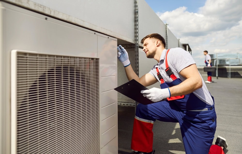 A maintenance worker in blue overalls and gloves inspects an outdoor HVAC unit on a rooftop. He holds a clipboard and appears to be taking notes. The sky is clear with some clouds in the background.