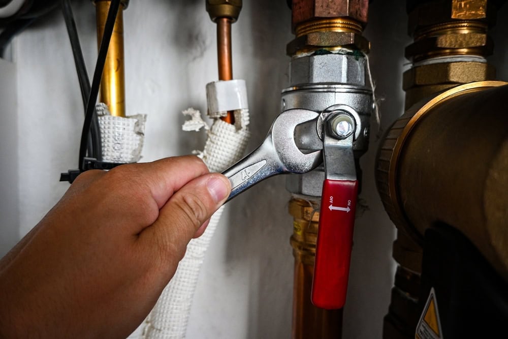 A hand grips a wrench to adjust a valve with a red handle in an industrial setting. Copper and metal pipes are visible, showcasing a plumbing or mechanical maintenance task.