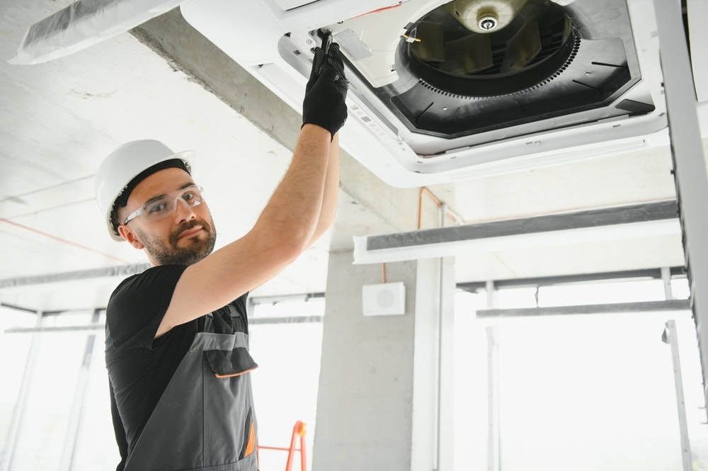 A construction worker wearing a white hard hat and black gloves is installing or repairing a ceiling air conditioning unit in a modern building. He is standing on a ladder and looking at the camera. The room has large windows and visible concrete structures.