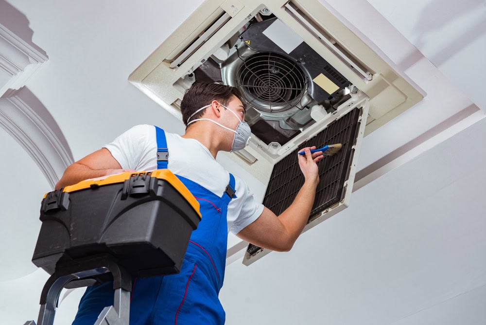 A person wearing a mask and blue overalls is standing on a ladder, working on a ceiling air conditioning unit. They hold a tool in one hand and a black toolkit in the other. The ceiling panel is open, exposing the unit.