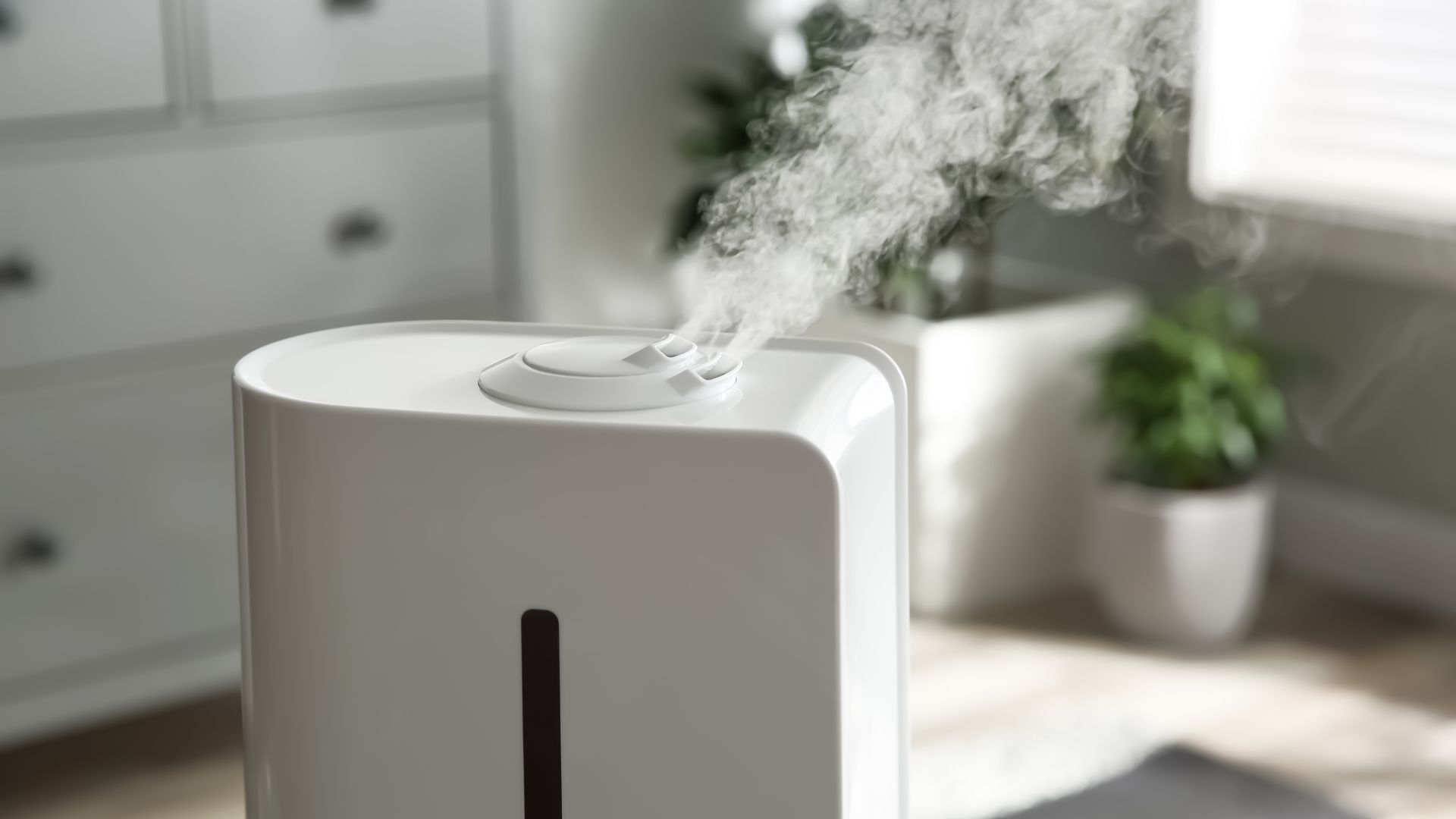 A white humidifier releasing a visible stream of mist in a bright room with green plants and white furniture in the background.