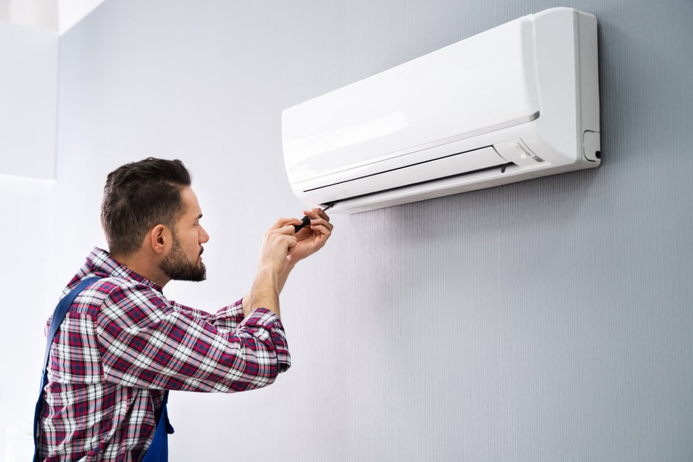 A man in a plaid shirt and overalls uses a screwdriver to repair or install a wall-mounted air conditioning unit, showcasing professional AC Services North Attleboro.