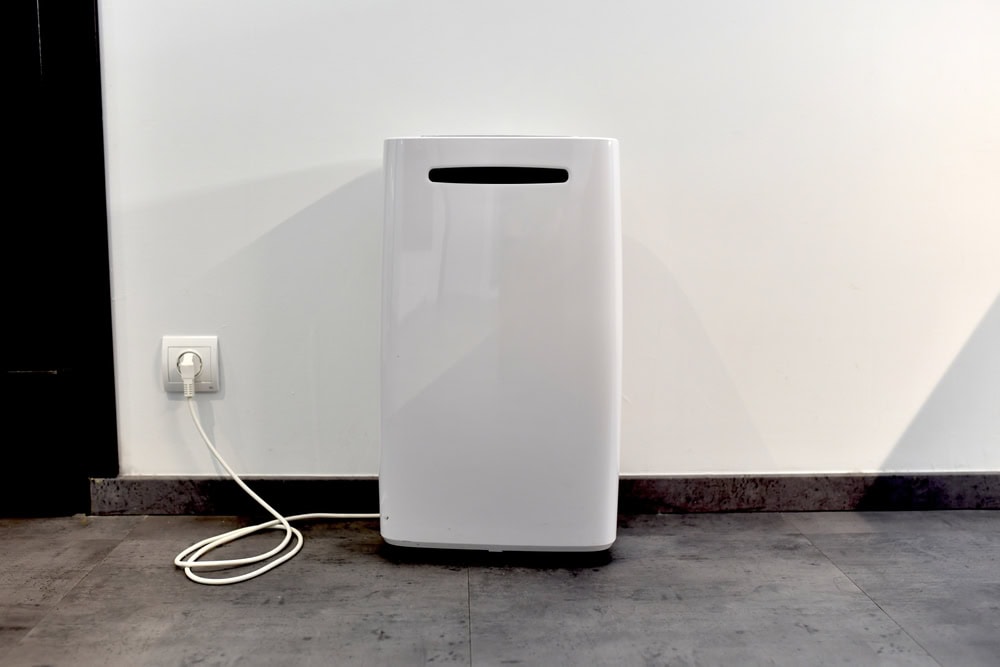 A white air purifier is plugged into a wall socket and placed on a gray tiled floor against a plain white wall.