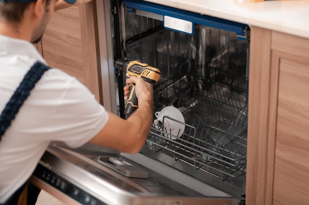 A person in work clothes uses a power drill to repair or install a partially open dishwasher in a kitchen.
