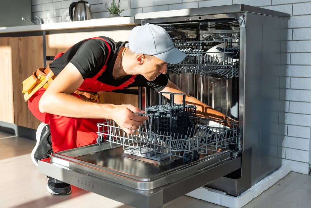 A person in a cap and red overalls kneels on the floor, inspecting or repairing an open dishwasher in a modern kitchen with wooden cabinets and sunlight streaming in.
