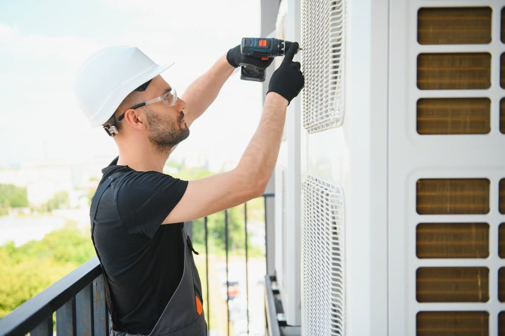A technician in a hard hat and safety glasses uses a power drill to work on a large outdoor air conditioning unit.