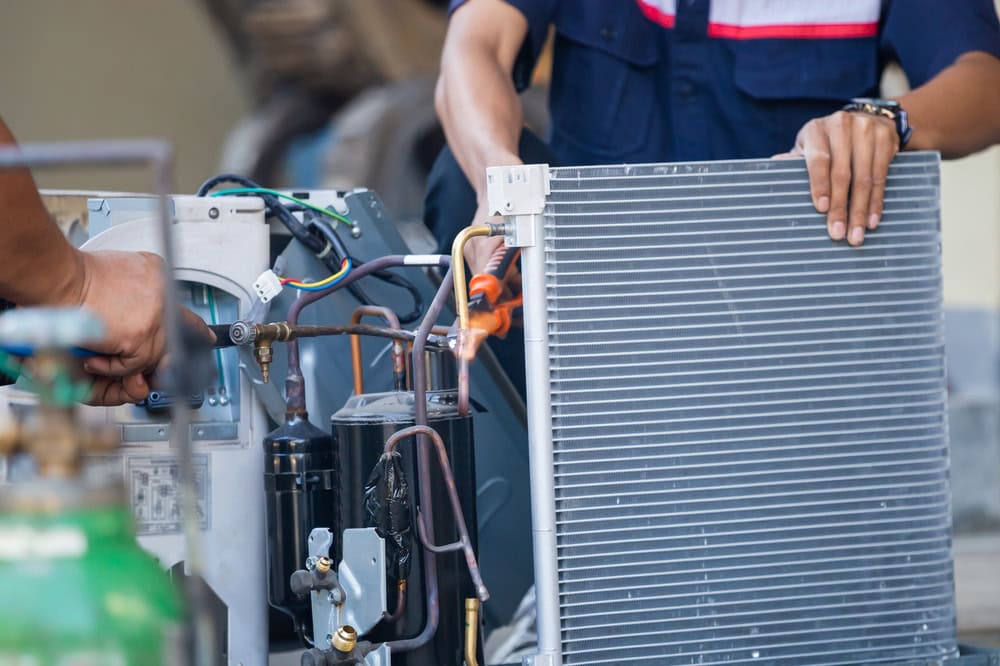 Two people repair an air conditioning unit, handling wires and a metal grille, with tools and machinery parts visible—showcasing the expertise of an HVAC Contractor North Attleboro, MA.