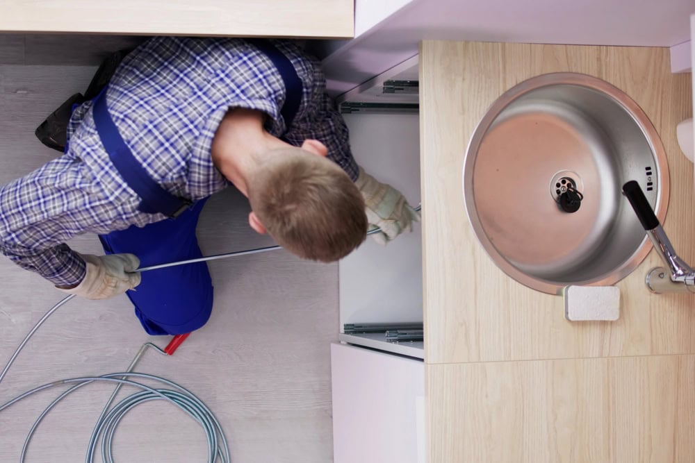A person in gloves and overalls uses a plumbing snake to unclog a pipe under a kitchen sink, viewed from above.