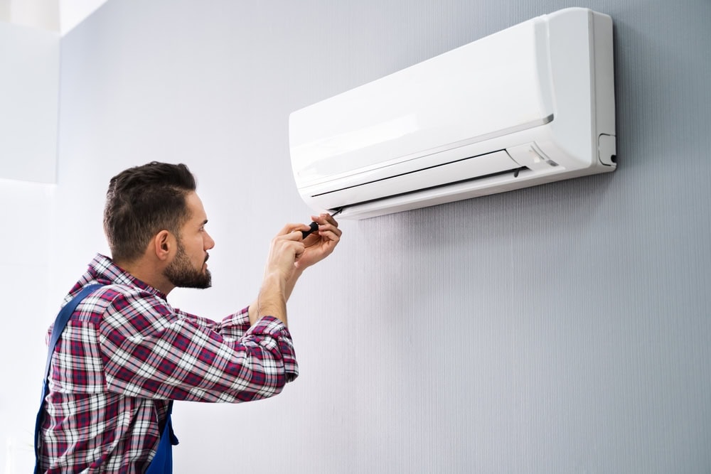 A man in a checkered shirt and overalls is repairing an air conditioning unit on a gray wall. Using his expertise as a skilled technician from MA, he's adjusting the AC with precision, fully focused on the task at hand.
