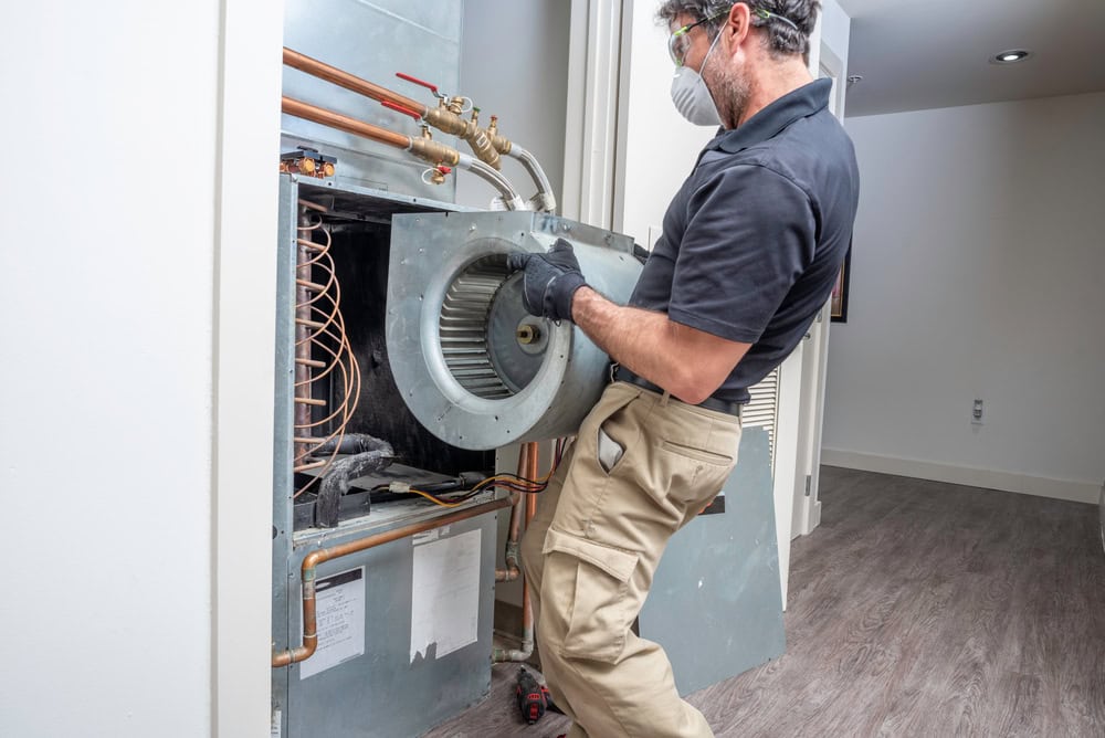 A person wearing protective gear, including goggles and a mask, is repairing a large HVAC unit. They are holding a cylindrical component inside the unit. The room has wooden flooring and a visible open doorway.