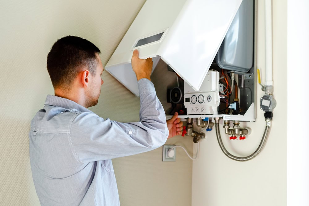 A man in a light blue shirt is inspecting or repairing a wall-mounted gas boiler, holding the cover open to access the internal components and controls.