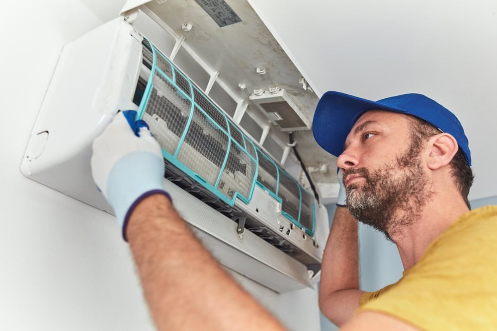 Person in a blue cap and yellow shirt repairing an air conditioning unit on the wall. They are wearing white gloves and are focused on adjusting the filter.