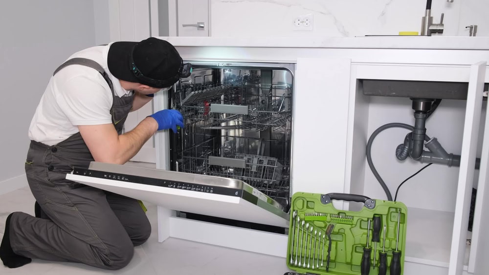 A technician wearing overalls and gloves kneels in front of an open dishwasher, inspecting it. A green toolbox with tools is on the floor nearby in a modern white kitchen.