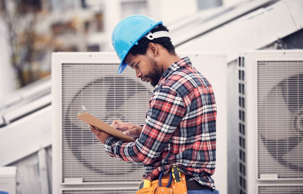 Worker in a blue hard hat and plaid shirt stands on a rooftop, holding a clipboard. He appears to be inspecting or taking notes near large air conditioning units. A tool belt is around his waist.