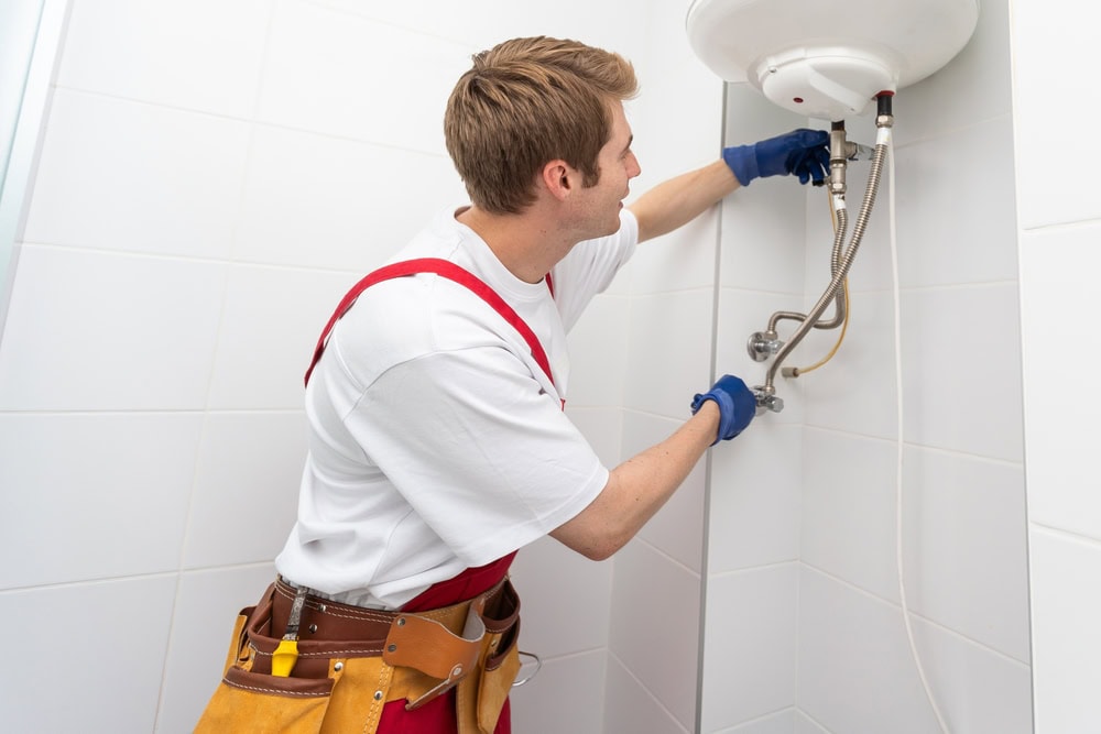 A plumber wearing a tool belt and gloves adjusts pipes connected to a water heater mounted on a white tiled wall.