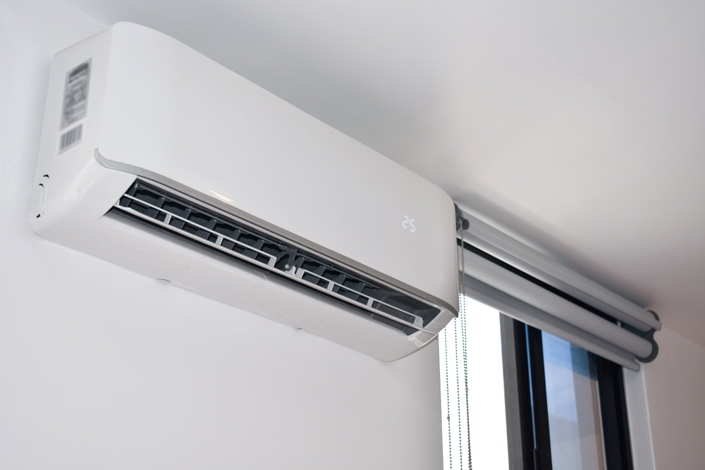 A white wall-mounted air conditioner is installed near the ceiling in a modern room, next to a window with a roller blind partially visible.