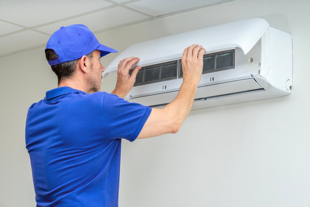 A technician in a blue uniform and cap inspects or repairs a wall-mounted air conditioning unit, lifting the front cover to access the internal components.