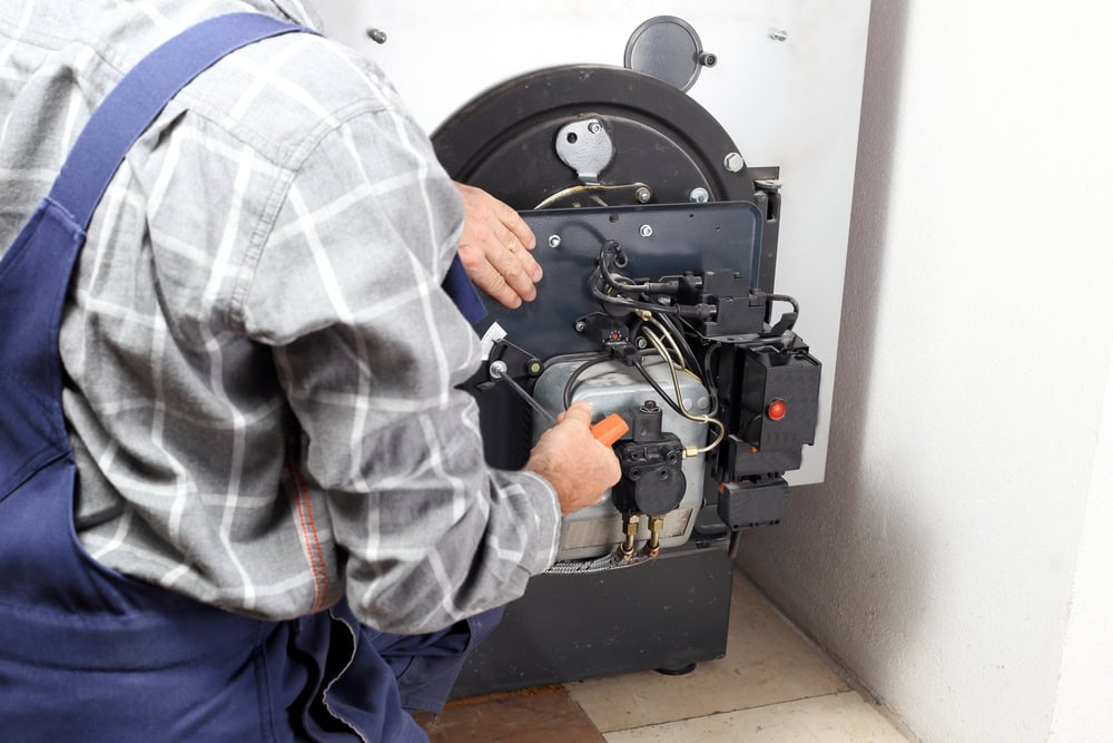 A technician in a plaid shirt and blue overalls is servicing or repairing a boiler or heating system, adjusting its controls and inspecting components.